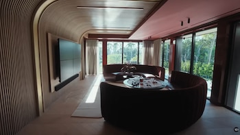Un salón moderno con paredes de lamas de madera curvas, sofás circulares rojos y una pantalla de video grande. Grandes ventanales muestran árboles