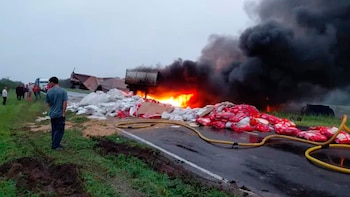 Camión en llamas con denso humo negro en carretera mojada. Bolsas de carga rojas y blancas esparcidas. Varias personas observan el accidente a la distancia