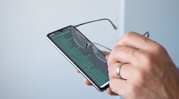 Una mano sostiene unos anteojos de marco metálico frente a un teléfono celular con letras verdes en la pantalla.