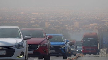 Vista frontal de una fila de automóviles y un camión en una carretera, con una ciudad difusa y cubierta de smog de fondo, indicando alta contaminación.