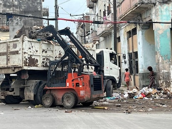 Miniexcavadora Toyota roja y negra junto a un camión de volteo blanco con basura. Dos mujeres, edificios deteriorados y cables eléctricos en la calle