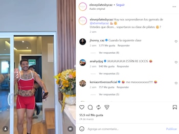 Eduin Caz, con bigote y tatuajes, sonríe a cámara, vestido con un conjunto deportivo rojo y sosteniendo un teléfono, en la entrada de un estudio con flores