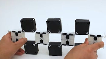 Dos manos sostienen una estructura horizontal compuesta por bloques cúbicos negros y piezas articuladas blancas sobre fondo blanco