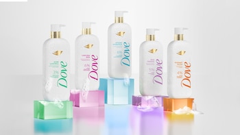 Cinco botellas blancas de jabón líquido Dove con dosificador dorado, sobre bloques rectangulares translúcidos de colores, con gotas de agua y espuma