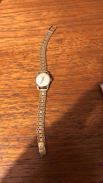Un reloj de pulsera dorado con esfera blanca y números oscuros, y una correa de eslabones, yace sobre una superficie de madera veteada de color marrón claro