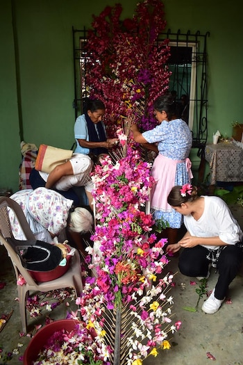 Varias mujeres, de espaldas y agachadas, colocan flores artificiales rosas, moradas, amarillas y blancas en una gran estructura vertical en una habitación verde