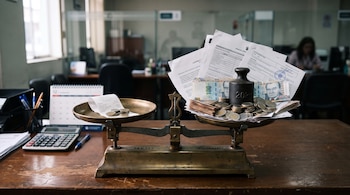 Balanza de metal sobre mesa de madera con billetes de 100 soles, monedas, peso de hierro, papeles, calculadora y calendario de fondo.