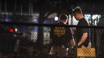 Dos agentes de la Unidad de Escena del Crimen de la NYPD son vistos a través de una valla metálica en una escena nocturna. Uno lleva una camiseta con "N.Y.P.D. POLICE CRIME SCENE UNIT"