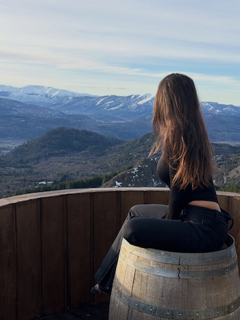 Una mujer vestida de negro está sentada sobre un barril de madera en un mirador frente a montañas nevadas