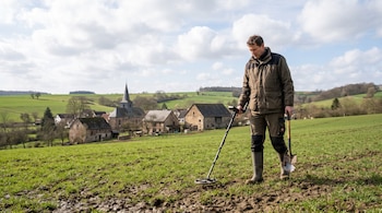 Hombre con abrigo, botas, un detector de metales y una pala en un terreno de hierba frente a un pueblo con iglesia.