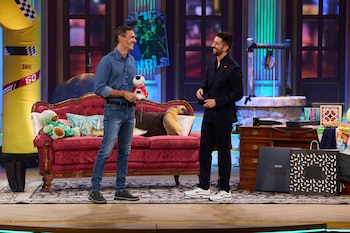Pedro Sánchez y David Broncano de pie en un plató de televisión frente a un sofá rojo, varios cojines y un escritorio de madera