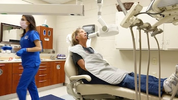 Mujer con bata protectora en sillón dental, con máquina de rayos X bucal. Una enfermera con uniforme azul y mascarilla camina al fondo de la clínica