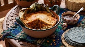 Cazuela de barro con un tamal en hojas de plátano, acompañada por un recipiente con salsa, tortillas azules y una calavera artesanal.