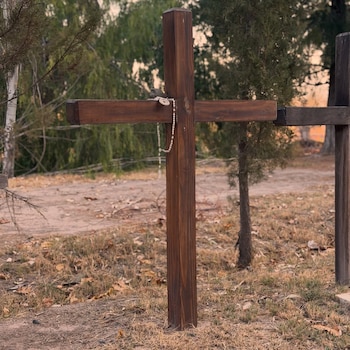 Una cruz de madera oscura con un rosario blanco colgando, en un campo de hierba seca y árboles al fondo durante el atardecer