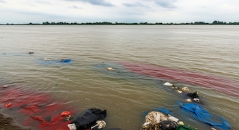 Río con orilla y aguas turbias marrones, bajo cielo nublado. Múltiples bolsas de basura, plásticos rojos y azules flotan cerca de la ribera con vegetación.
