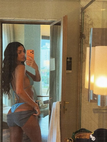 Zaira Nara de espalda, tomando una selfie en el espejo con un teléfono naranja, vistiendo un bikini celeste y bajando unos shorts grises