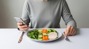 Primer plano de una persona en camiseta gris consultando su móvil junto a un plato con espinacas, pepino, zanahorias, brócoli y pollo; un cuchillo y un tenedor están sobre la mesa.