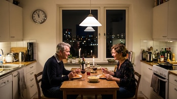 Un hombre y una mujer de mediana edad cenan pasta con vino tinto en una mesa de madera. Hay velas, un reloj a las 23:00 y un ventanal nocturno.