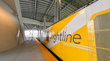 Vista lateral de un tren Brightline blanco y amarillo en una estación moderna con techo metálico, pilares de hormigón y plataforma con franja táctil amarilla