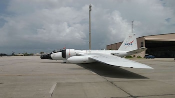 Avión blanco y negro con la insignia de la NASA en el estabilizador vertical sobre una pista de cemento. Se observan un hangar, un poste y un helicóptero