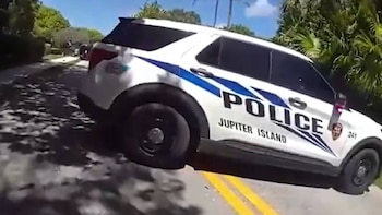 Un SUV policial blanco de Jupiter Island con franjas azules en una carretera. Se aprecian árboles verdes y un cielo azul claro de fondo