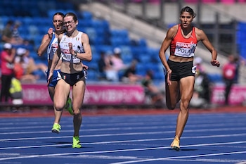 Tres atletas femeninas corren en una pista de atletismo azul. La del centro viste de blanco y negro, la de la derecha de rojo y negro, la de la izquierda de azul