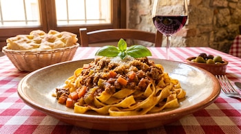 Un plato de pasta tagliatelle con salsa ragú, queso rallado y hojas de albahaca, acompañado de focaccia, vino tinto y olivas.