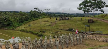 Soldados en uniformes de camuflaje de espaldas, formando en un terreno verde con un lago. Al fondo, banderas y más militares en una colina con selva y cielo nublado