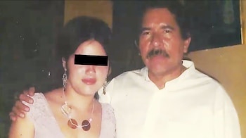 Un hombre y una mujer posan para la cámara. El hombre tiene bigote. La mujer tiene una barra negra que cubre sus ojos. El hombre la abraza