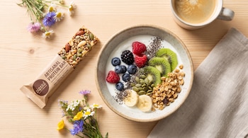 Barrita proteica, cuenco de yogur con frambuesas, arándanos, kiwi, plátano, semillas de chía y calabaza, granola, taza de café, flores y paño en mesa de madera.