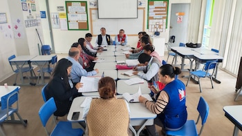 Un grupo de personas sentadas alrededor de varias mesas unidas en un salón de clases con pizarrón, sillas azules, y carteles informativos en las paredes