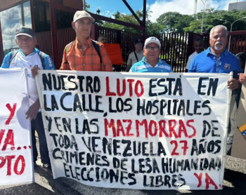 Varios hombres sostienen una pancarta blanca con texto en negro y rojo que dice: "NUESTRO LUTO ESTÁ EN LA CALLE, LOS HOSPITALES Y EN LAS MAZMORRAS DE TODA VENEZUELA 27 AÑOS CRÍMENES DE LESA HUMANIDAD ELECCIONES LIBRES YA"