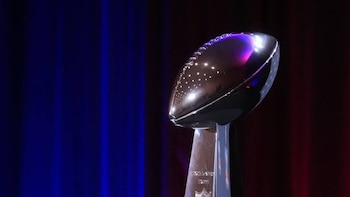 Un primer plano del trofeo Vince Lombardi, brillante y metálico, con forma de balón de fútbol americano sobre una base, iluminado por luces azules y rojas