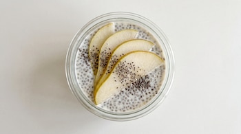 Vaso de cristal con pudín de chía y kéfir, rodajas de pera y semillas de chía en la superficie. Fondo claro y uniforme.