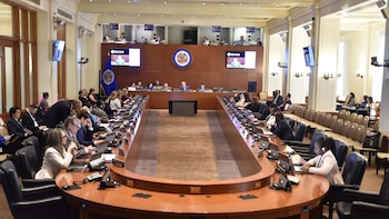 Sala de conferencias amplia con mesas en U, múltiples delegados con computadoras, pantallas mostrando presentaciones y el emblema de la OEA y su bandera