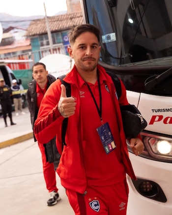 Hombre joven con barba y bigote, vestido con conjunto deportivo rojo y credencial, saluda con pulgar hacia arriba junto a un autobús