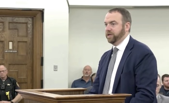 Un hombre con traje azul y corbata gris habla detrás de un podio de madera en la sala de un tribunal