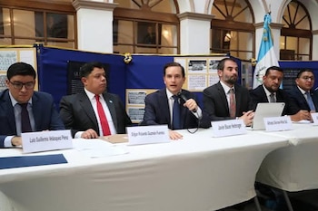 Seis hombres con traje están sentados ante una mesa blanca con carteles con sus nombres y la bandera de Guatemala al fondo