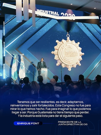 Un hombre con traje habla tras un podio en un escenario con banderas y una pantalla gigante que muestra una medalla