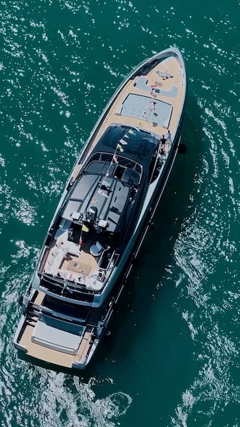 Vista aérea de un yate de lujo color negro y blanco con cubiertas de madera clara navegando en aguas azules y brillantes bajo el sol