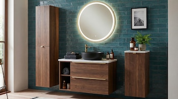 Vista de un baño con pared de azulejos verde azulado, un tocador de madera oscura con lavamanos de cuenco negro, un espejo redondo iluminado y gabinetes flotantes.