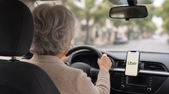 Vista posterior de una mujer de cabello cano al volante de un automóvil con un teléfono que muestra la pantalla de Uber.