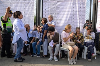 Varias personas, algunas sentadas y otras de pie, observan grandes paneles blancos con listas de nombres y mesas de sufragio en un centro de votación en Lima
