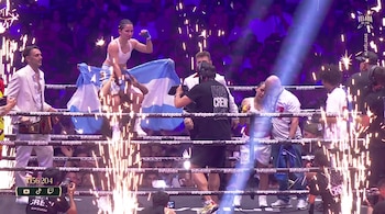 Boxeadora alzada en hombros con una bandera argentina en un ring con fuegos artificiales, rodeada de personas y público en un evento.