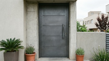 Puerta de entrada de acero gris oscuro con cuatro paneles horizontales y manija larga. Flanqueada por paredes de hormigón y estuco claro, con plantas en macetas.