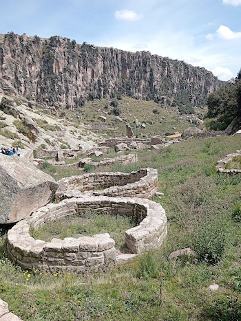 Vista panorámica de las ruinas de Taqrachullo, mostrando muros de piedra circulares entre vegetación y rocas, con una gran pared rocosa de fondo bajo un cielo azul con nubes