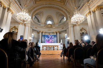 Salón ornamentado con columnas doradas, dos candelabros de cristal, un escenario, una mesa, una pantalla digital, dos banderas y personas sentadas