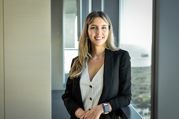 Mujer con cabello castaño claro, blazer negro y blusa blanca sonríe frente a una ventana. Lleva un collar, pulseras y reloj inteligente
