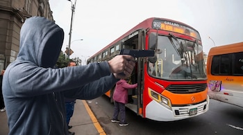 Hombre encapuchado con buzo gris apunta una pistola negra a un autobús rojo de pasajeros. Un segundo autobús naranja está a la derecha, personas en la calle.