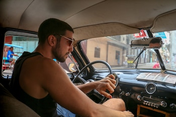 Hombre barbudo con gafas de sol, camiseta sin mangas, conduciendo un coche clásico. Interior del vehículo con volante, salpicadero y calle visible por el parabrisas